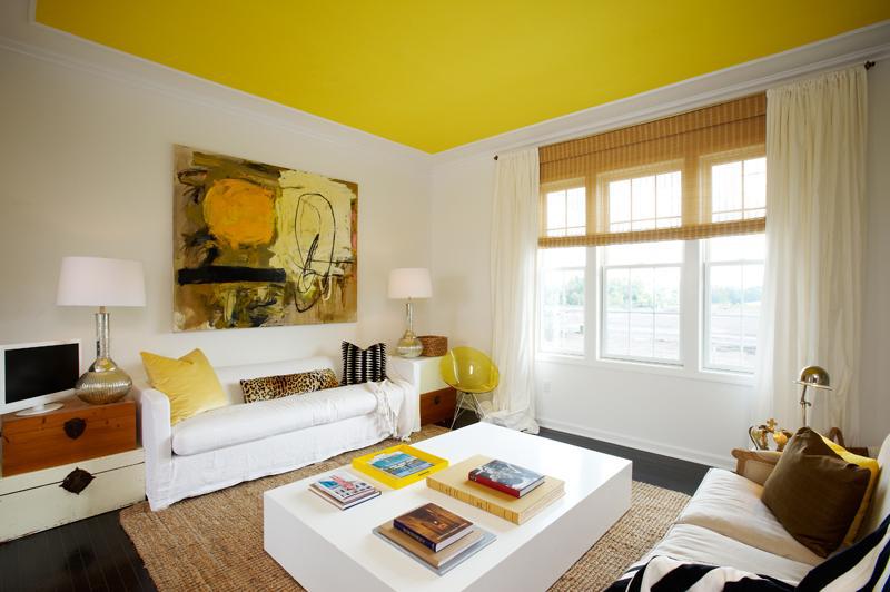 Teto colorido na sala: amarelão compondo com as almofadas, quadro, cadeira e elementos decorativos do ambiente. Paredes e móveis brancos.